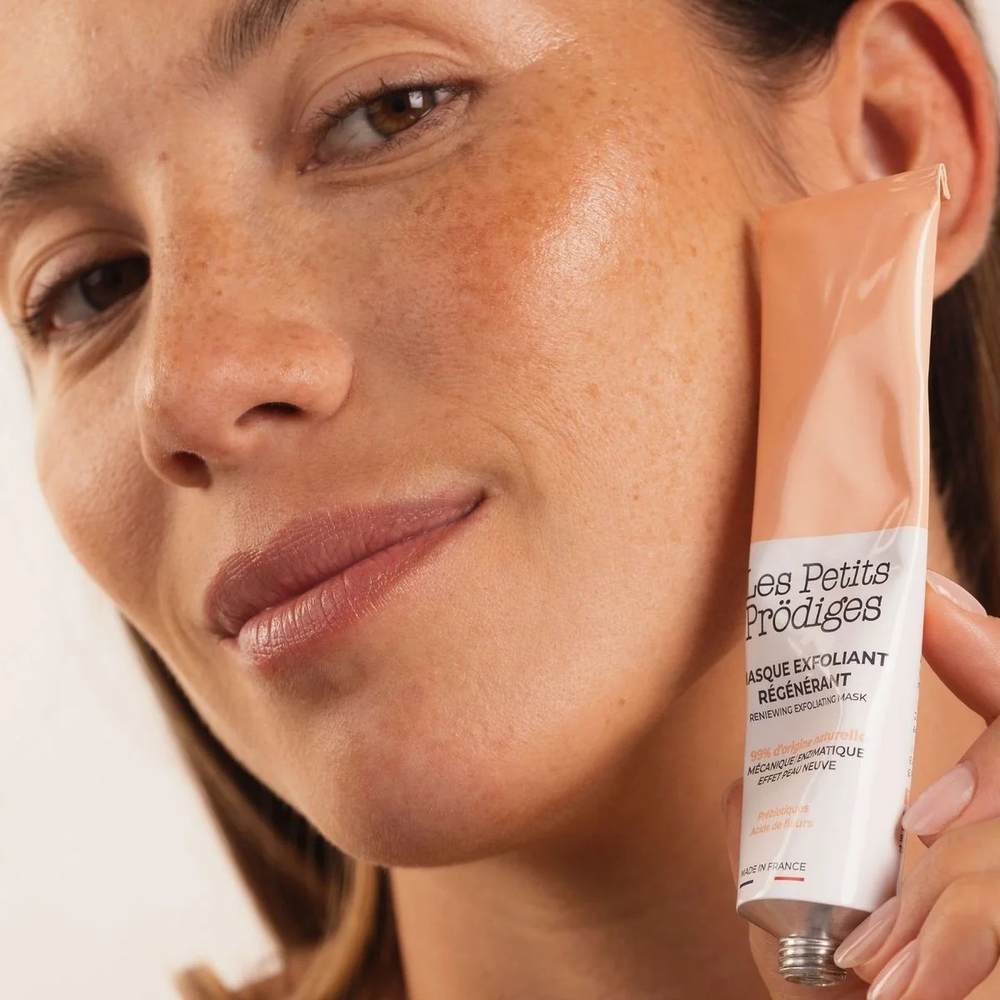 Femme au teint lumineux tenant le tube du masque exfoliant régénérant contre sa joue